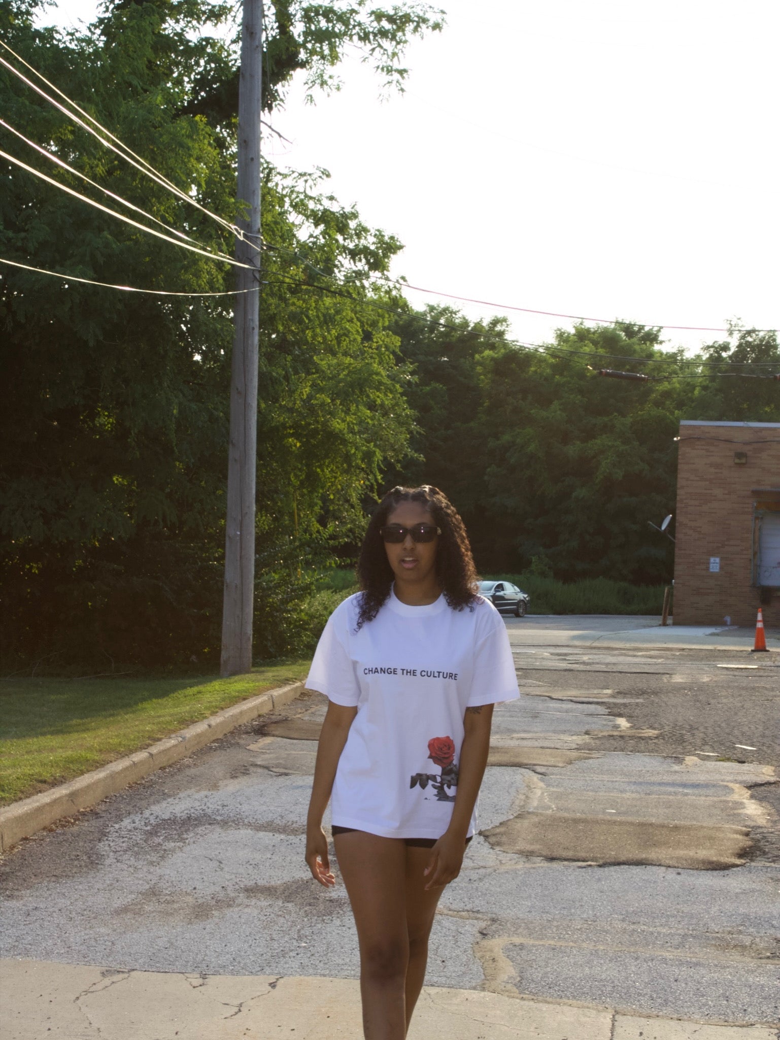 Person wearing a white t-shirt with graphics on a street with trees and a building in the background