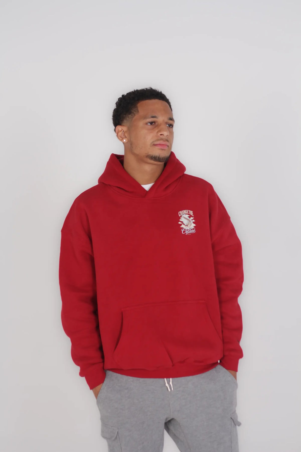 A man wearing a red pullover hoodie with a small white graphic on the left chest, paired with gray sweatpants, standing against a plain light gray background.