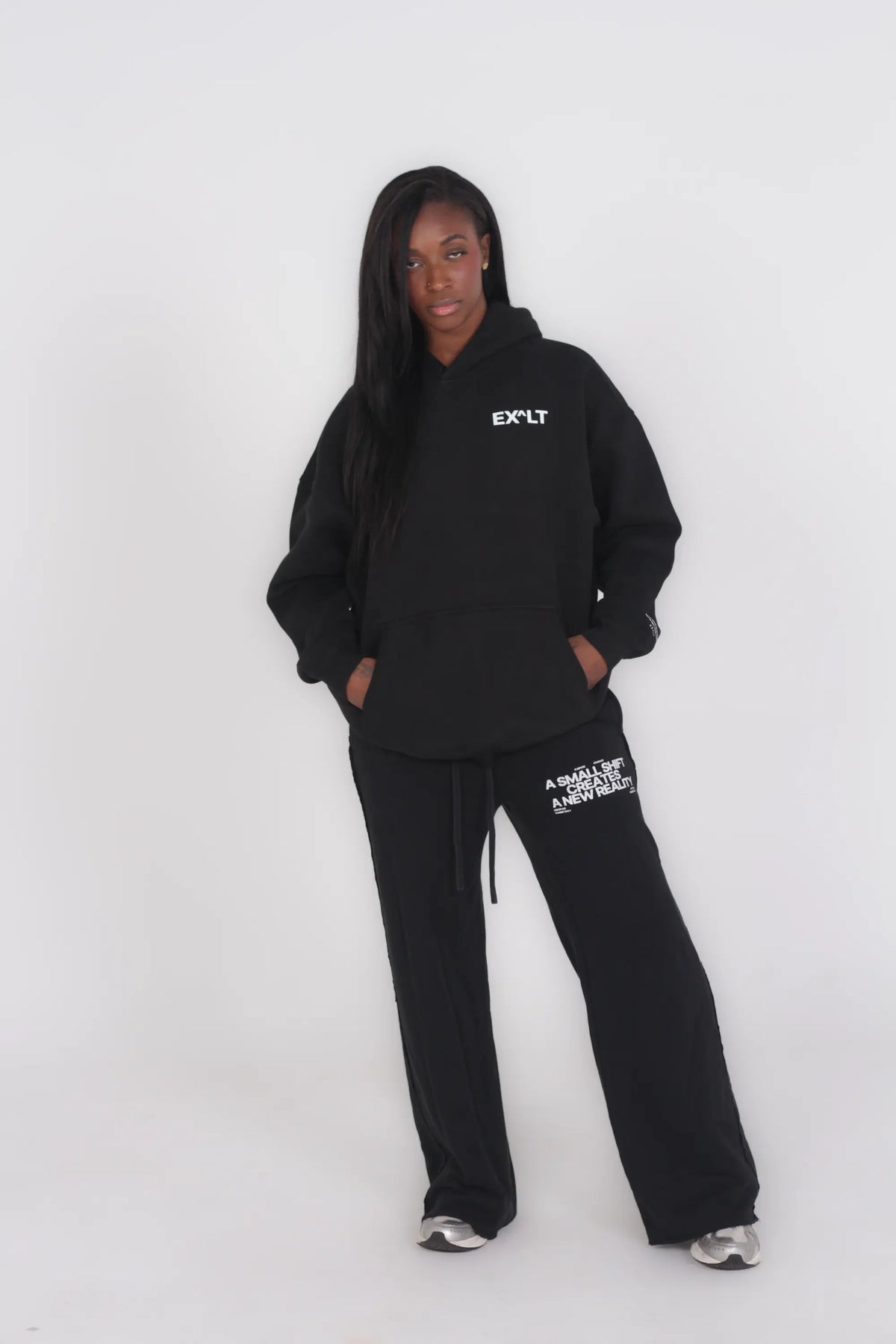 A model wearing a black hoodie and matching sweatpants with white text graphics, standing against a plain light gray studio background.